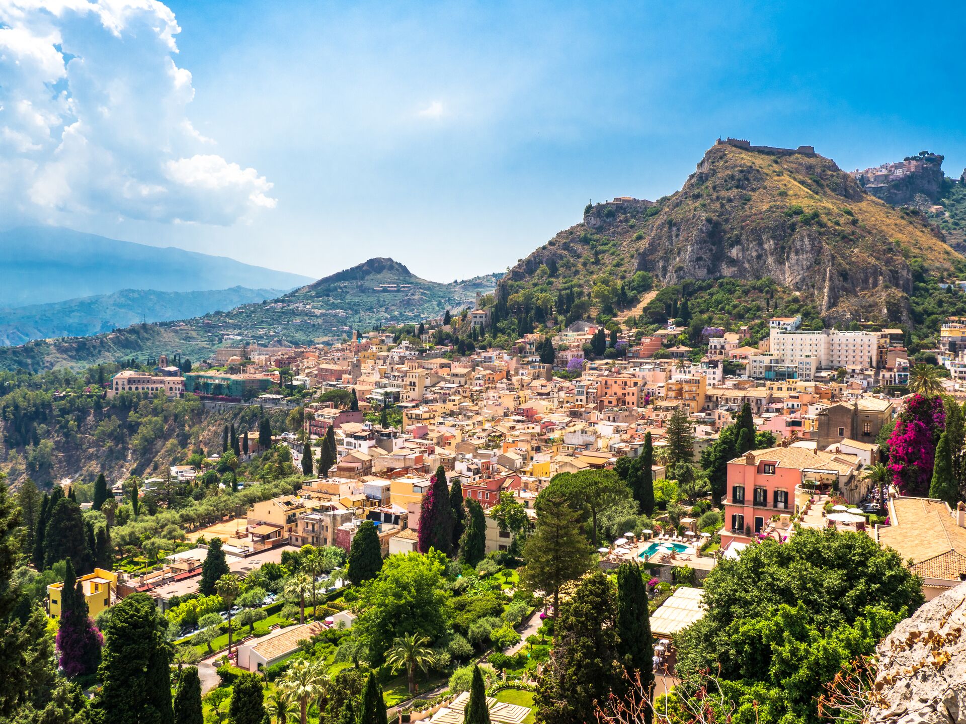 View across the golden houses of Taormina, with a blue sky and Mount Etna behind, and green trees in the foreground