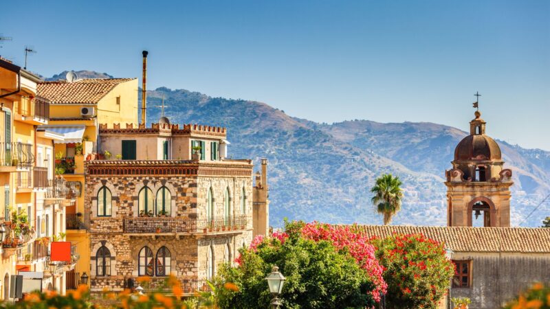 Sunshine view over Taormina with yellow buildings in front and mountains behind, showing the spectacular architecture