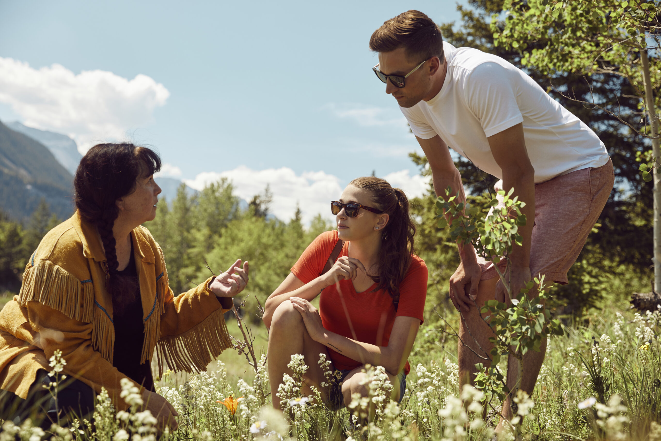 Brenda, crouched down, shows plants to two guests, with white flowers in front and a bright blue sky and sunshine