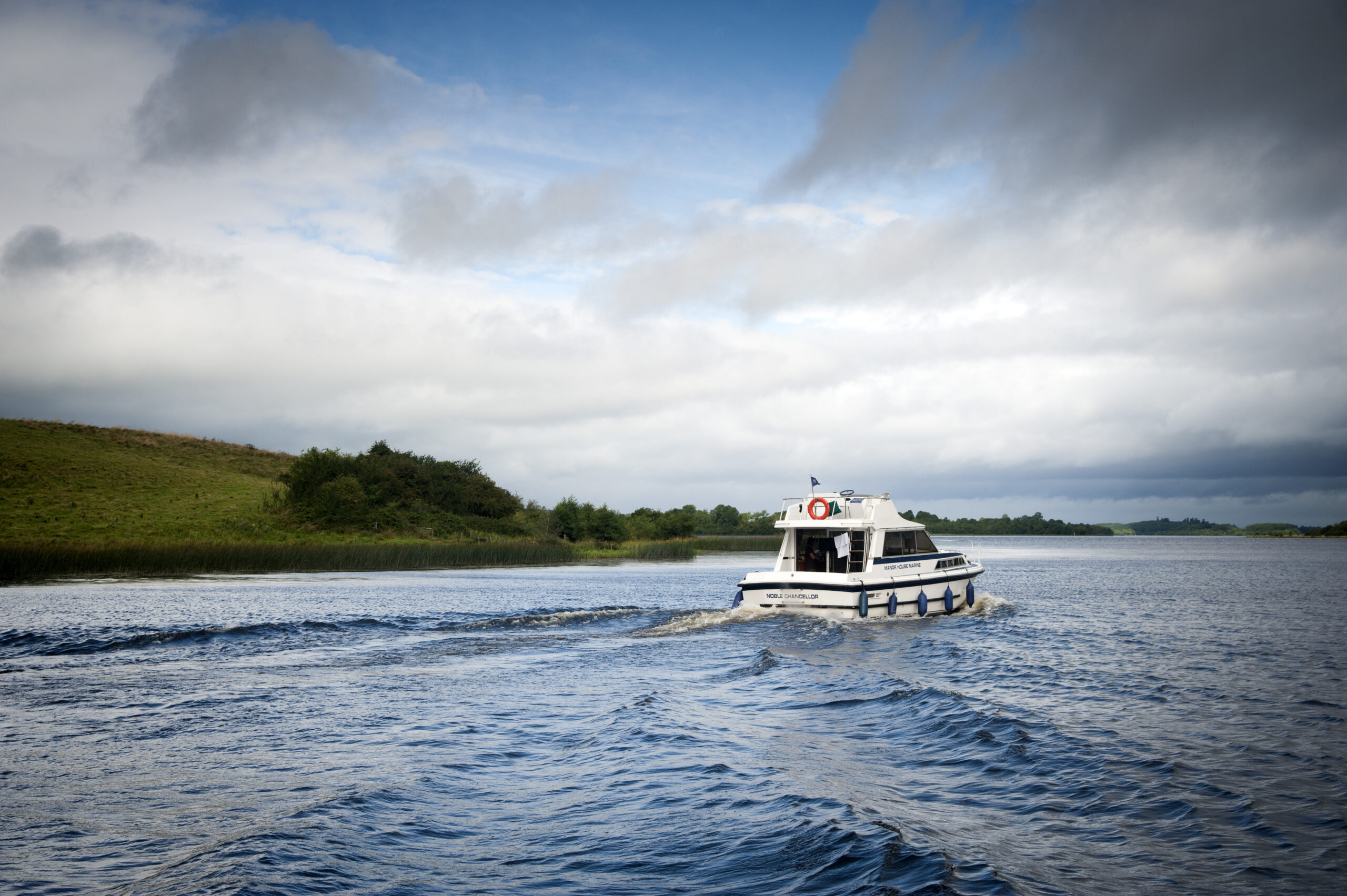 A white boat sails across the lake waters, against a cloudy sky and green hills in the background