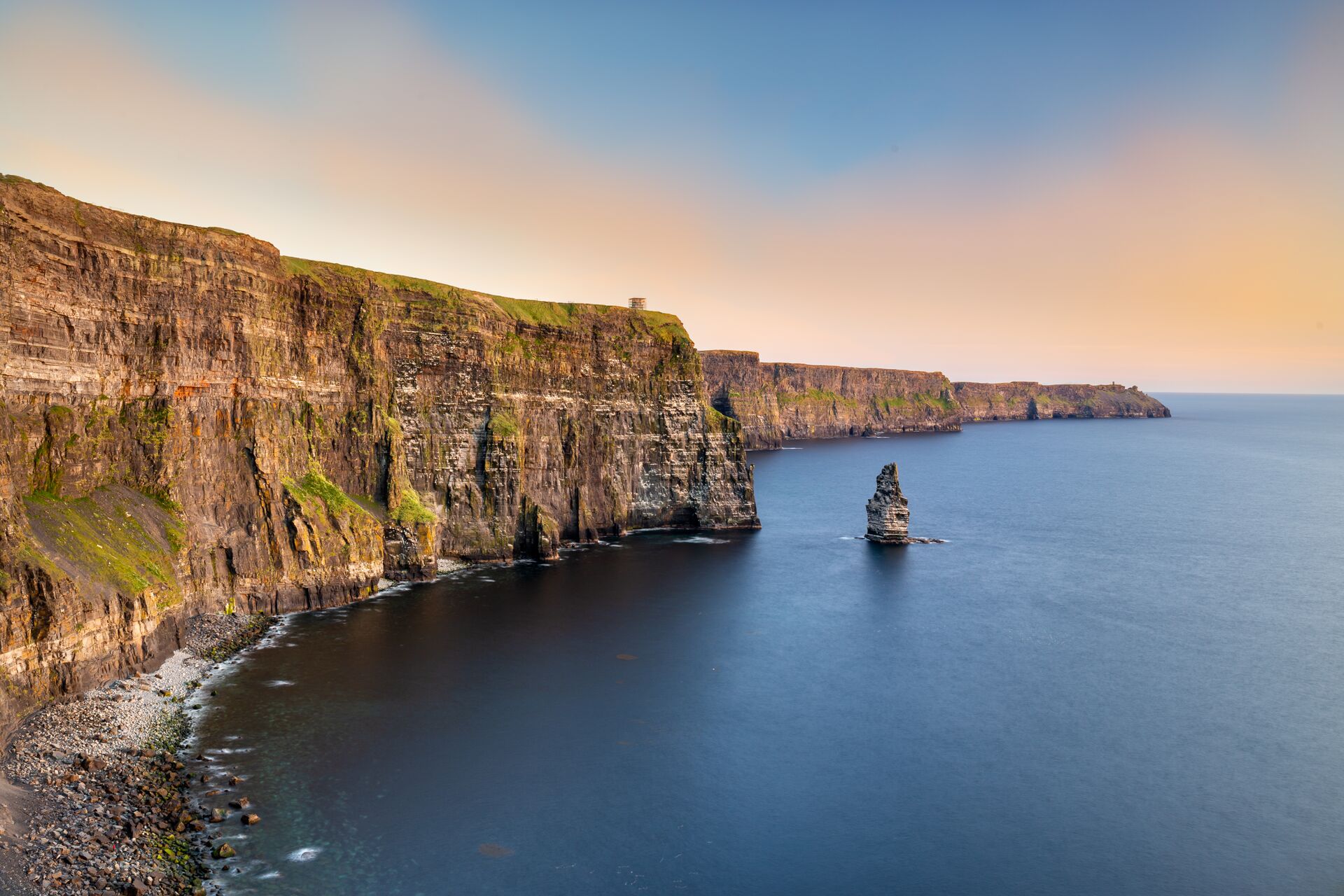 The striking high cliffs of Moher overlook the dark blue waters with a blue sky and lighlty coloured clouds up above