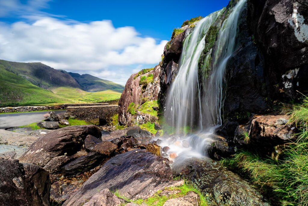 a hige waterfall stands with the Wild Atlantic Way road in the background, with brown rocks and bright green foliage and a bright blue sky with white clouds