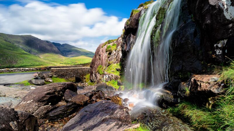 a hige waterfall stands with the Wild Atlantic Way road in the background, with brown rocks and bright green foliage and a bright blue sky with white clouds