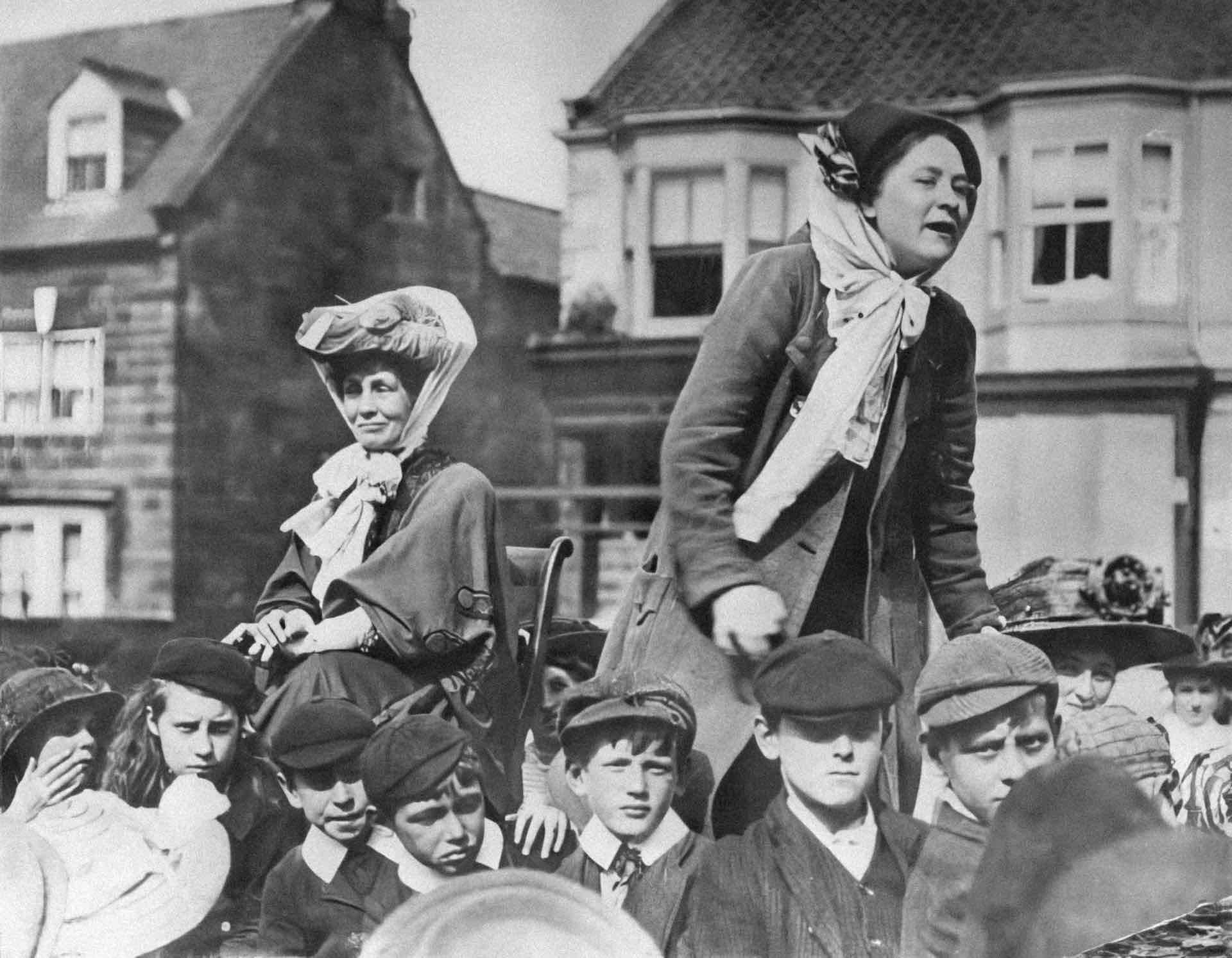 Black and white image of two women being carried on chairs above a crowd 