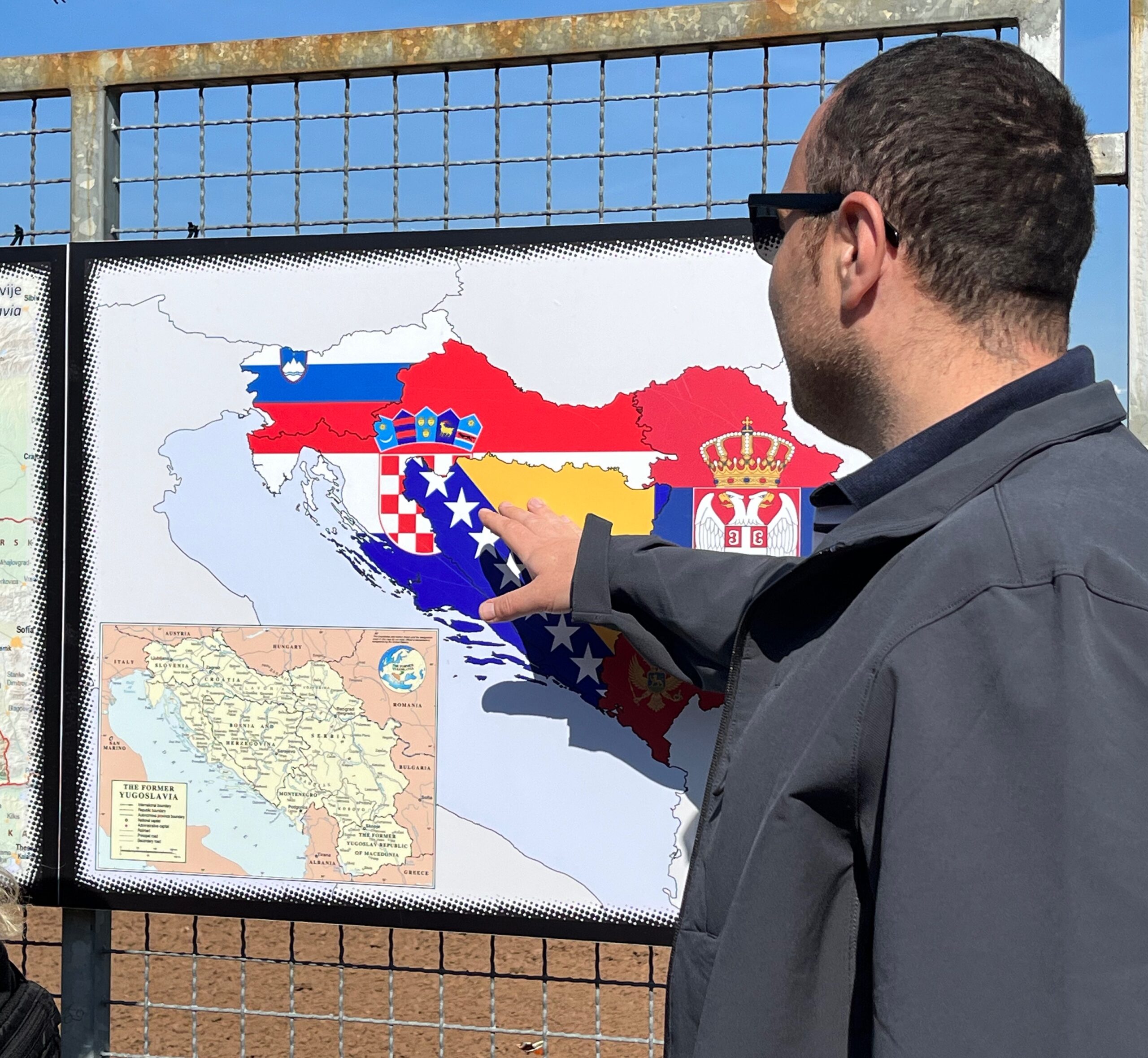 Aldin is pictured in a blue jacket and sunglasses, pointing to a map of the Sarajevo war tunnel in the sunshine