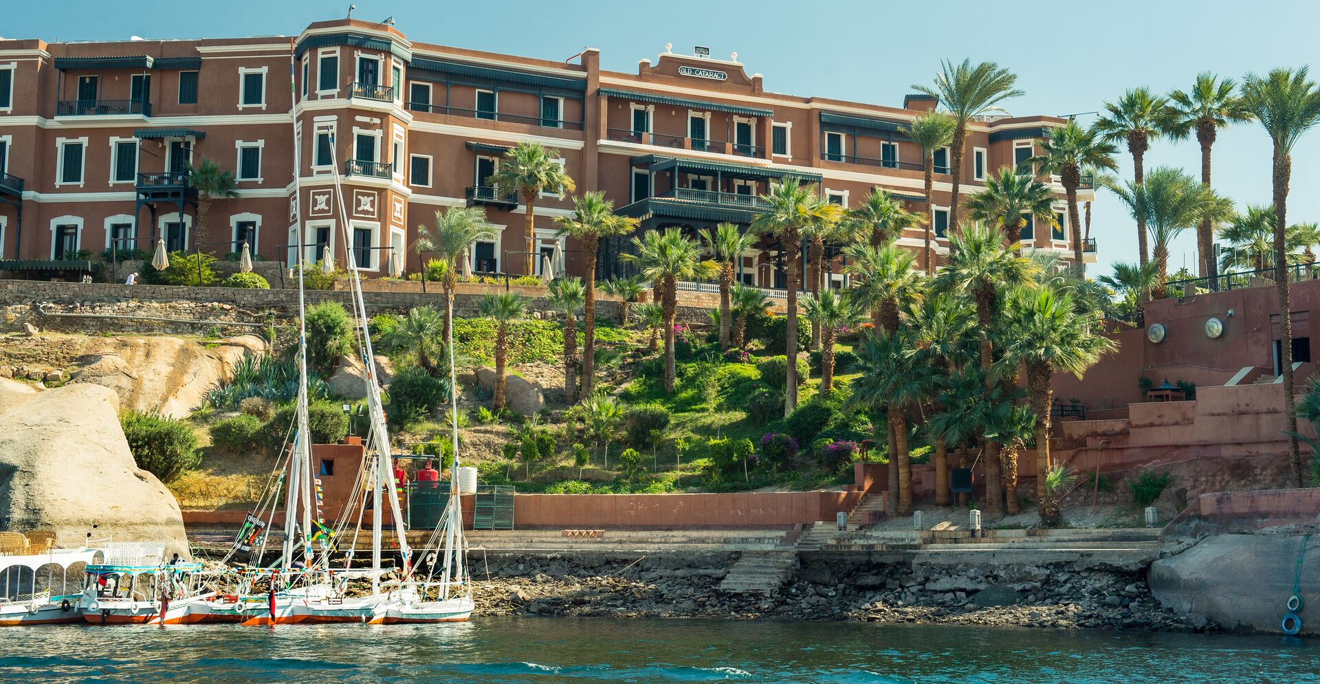 The Old Cataract Hotel is pictured with green trees and the River Nile in front