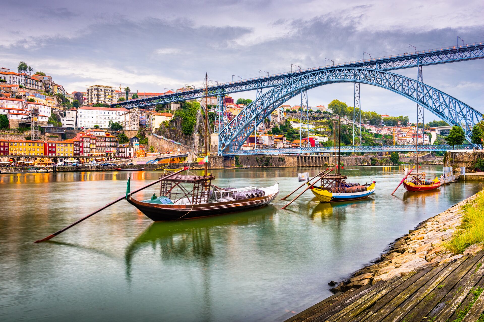 Boats in bright primary colours sit on the River Duoro in Porto, with a right blue bridge behind and colourful houses climbing the hillside in the background