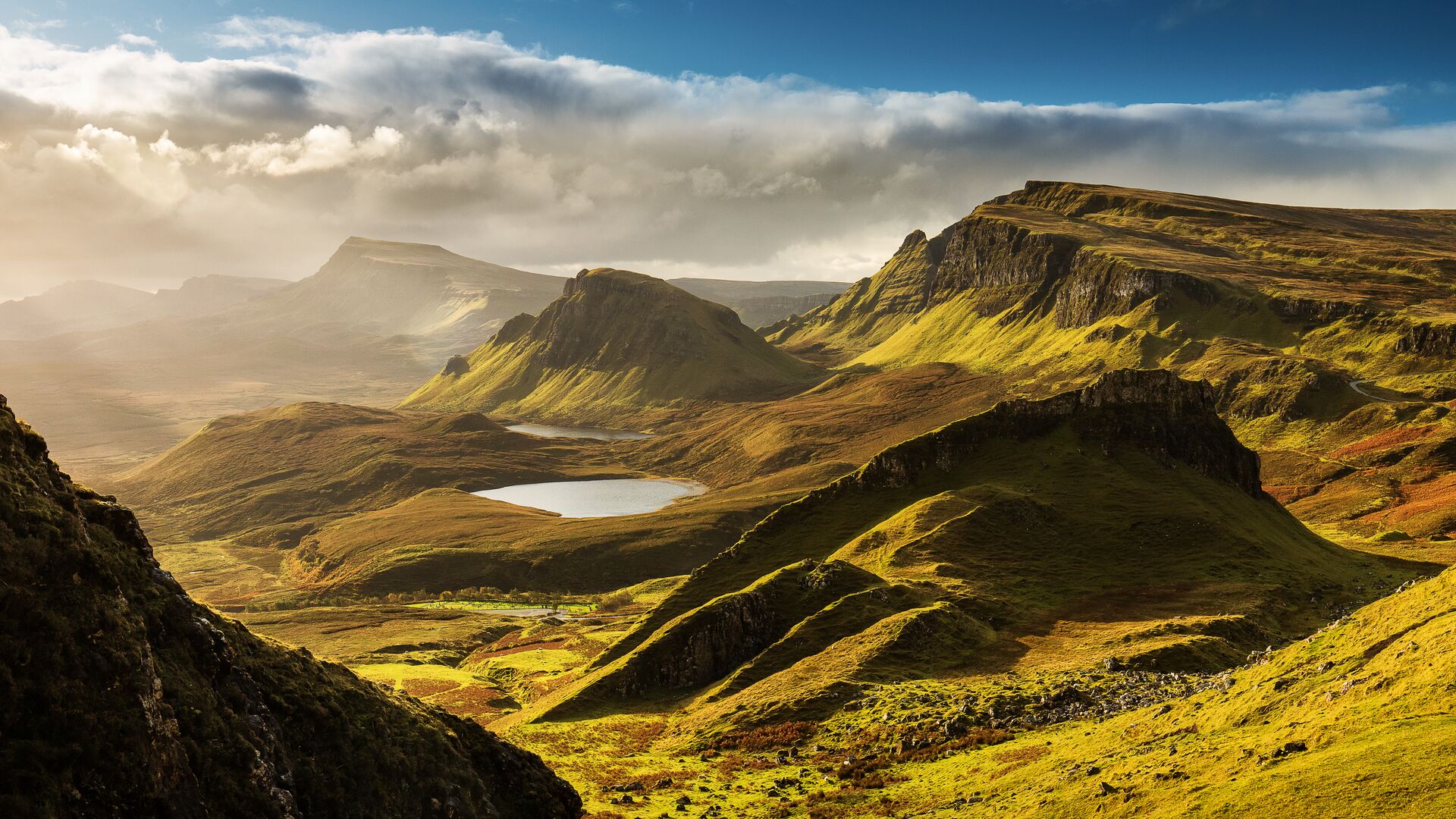 Green mountains and valleys in Scotland shine in the sun with mist in the background and a bright blue sky