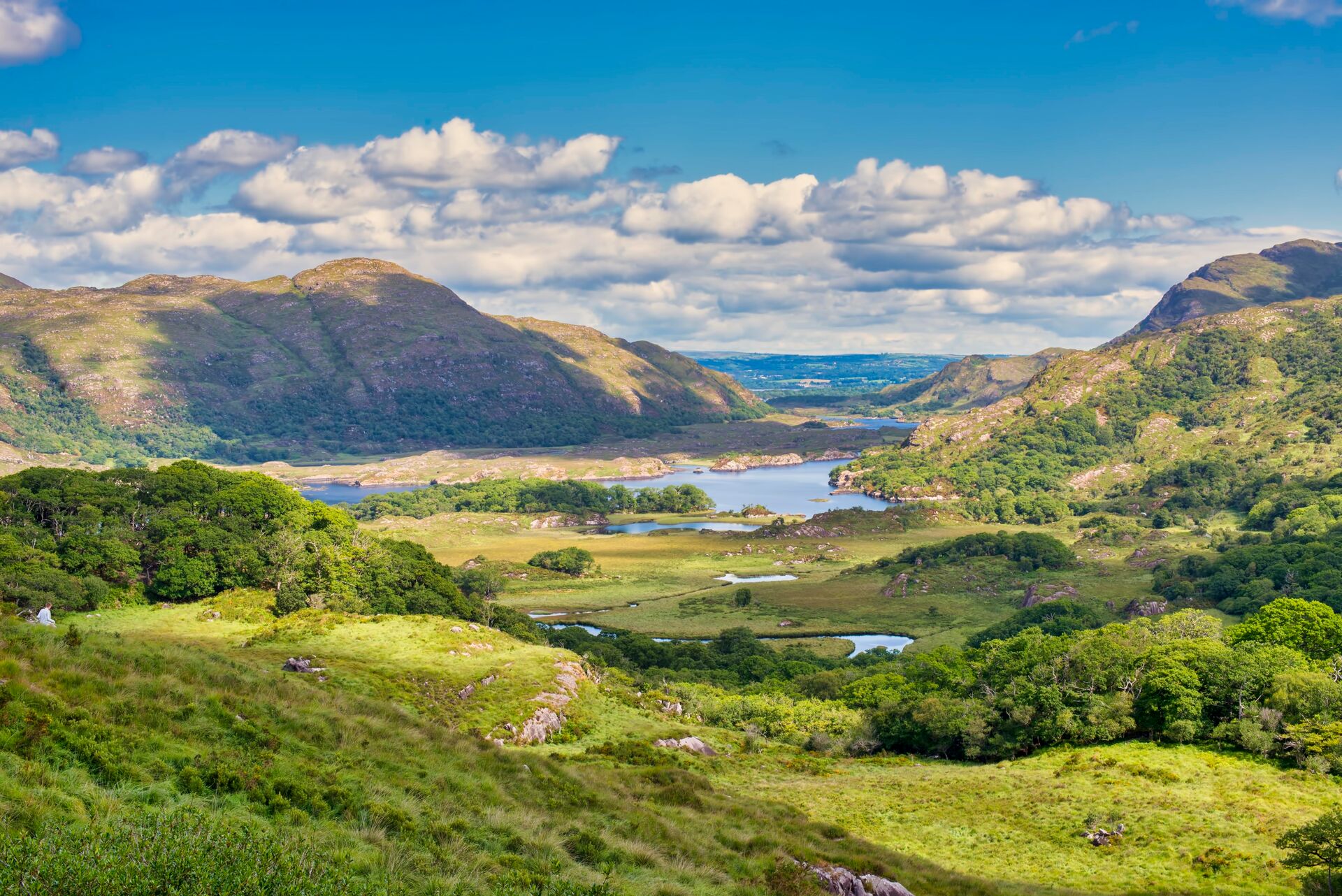 Green fields and valleys fill the image, with blue sea and mountains behind, against a backdrop of a blue sky with white fluffy clouds.