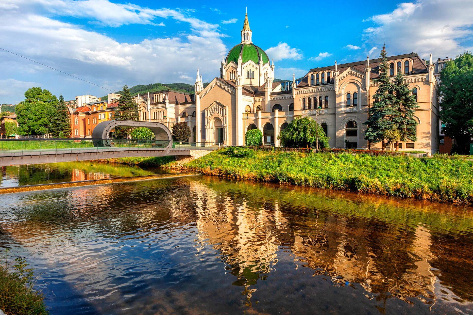 An ornate white building with a green roof in the historic centre of Sarajevo, with a river flowing in front