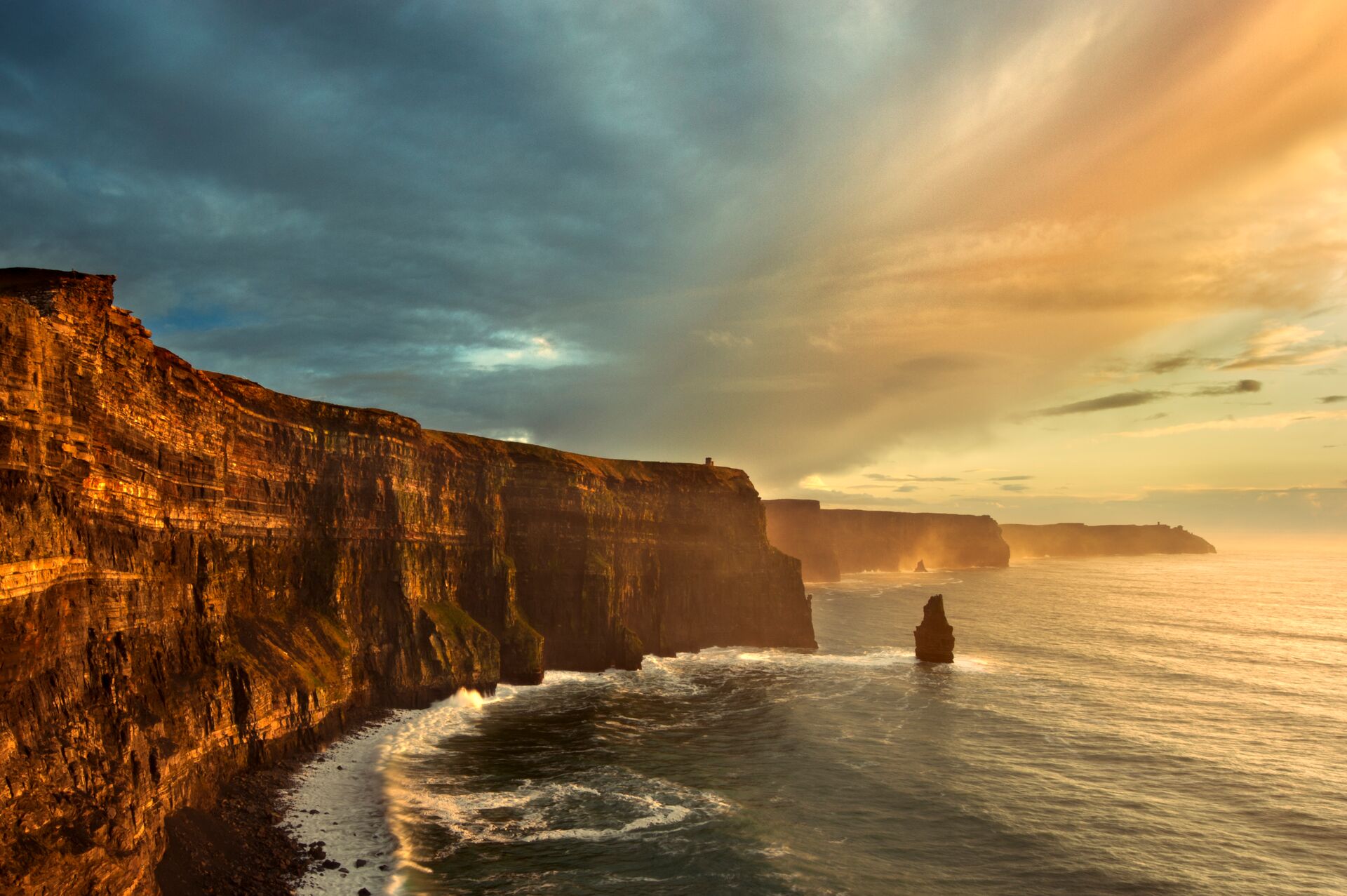 Waves crash against tall rock cliffs, with a moody sky of blue, yellow and clouds