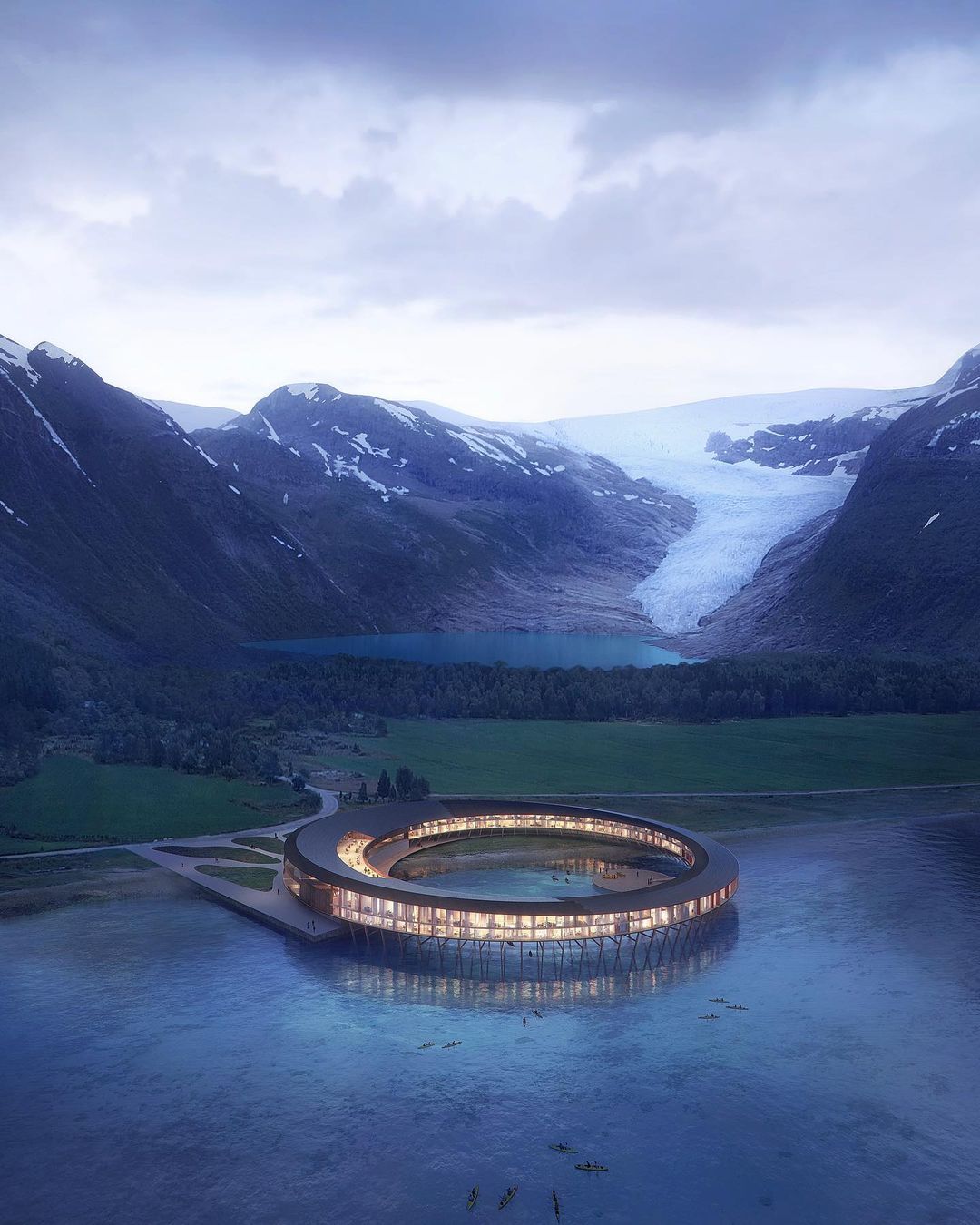 The circular Svart Hotel sits on a glacier lake, with light on inside and grey atmospheric skies and mountains in the background