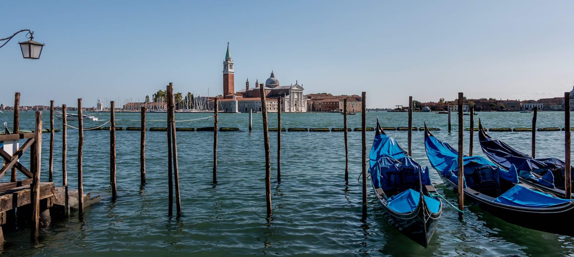 Two gondolas are moored up by a pier in Venice, water stretches out towards old historical buildings in the distance.
