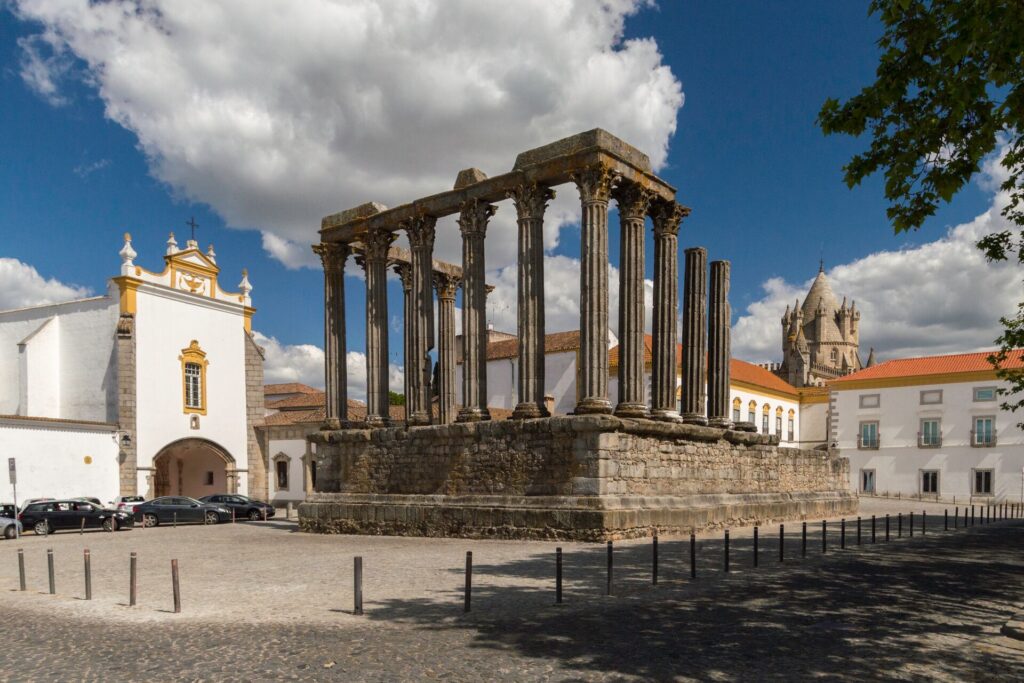 An ancient Roman Temple with many dark stone columns sits int he middle of a cobblestone square, with white buildings and red roofs in the background.