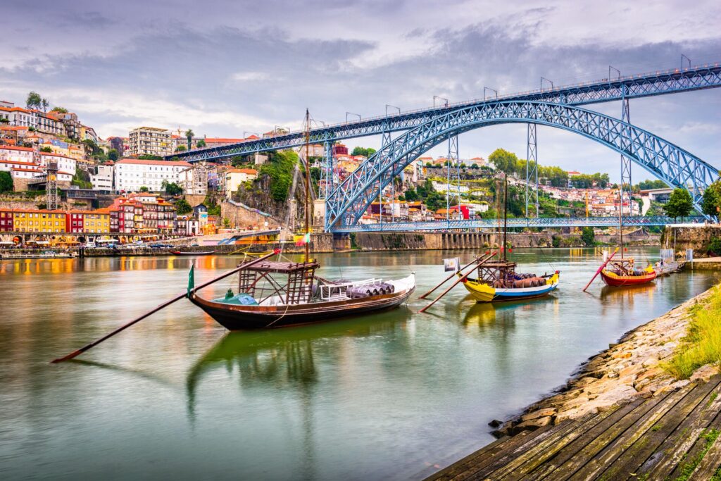 Traditional boats glide on the Douro River in Porto, with the colourful houses behind and a large iron bridge spanning the tow banks.