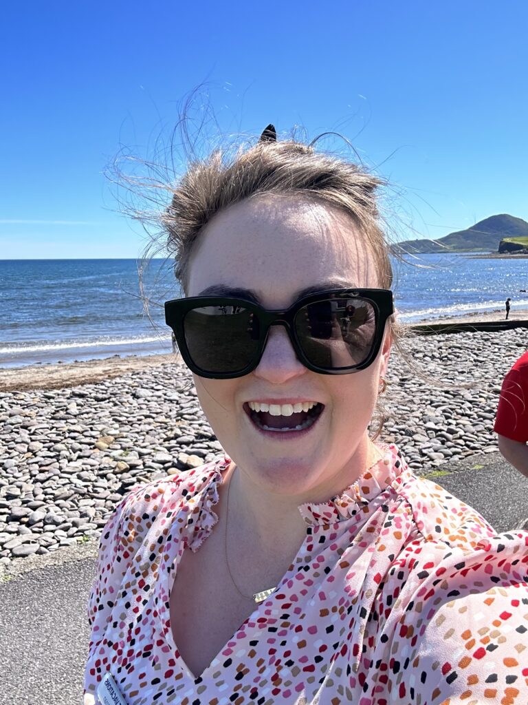 A picture of Faith, one of our global travel experts, taking a selfie at a beach with sunglasses on and a bright blue sky in the background.