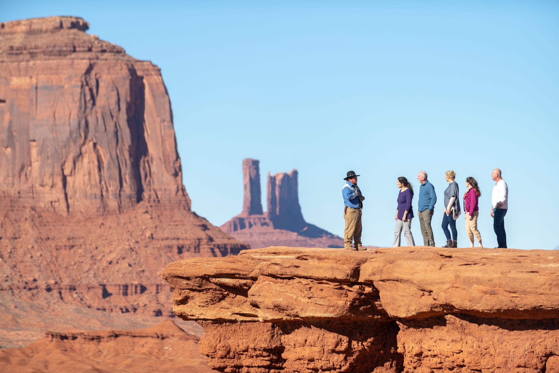 Photo of Larry standing on John Ford Point at Monument Valley, talking to some guests. Shot from distance, with monument valley buttes in the background