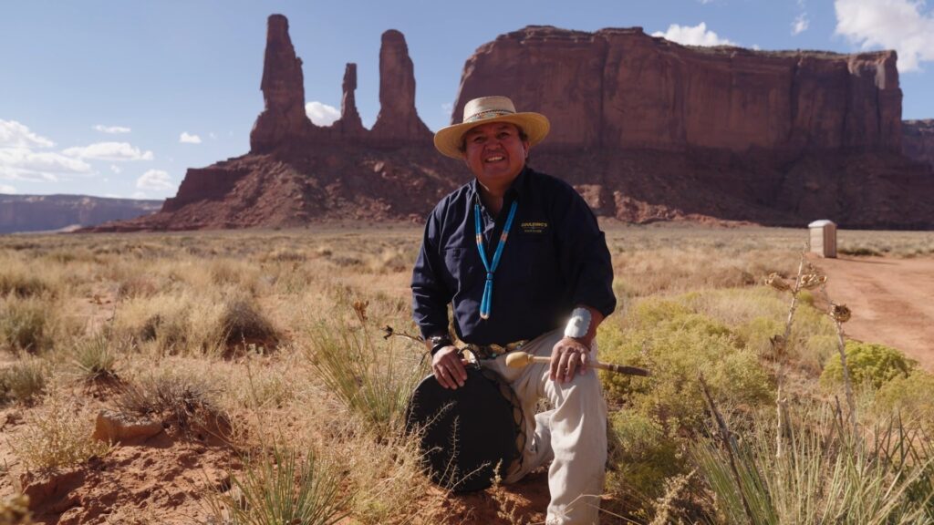 Portrait photo of Navajo guide Larry Holiday kneeling in front of the Three Sisters rock formation in Monument Valley
