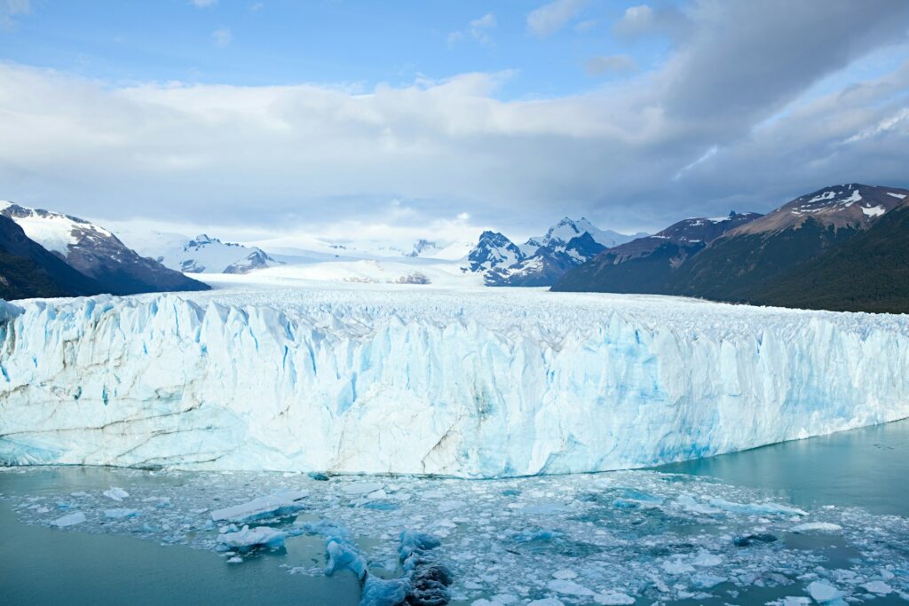 Image of a large glacier, taken from a distance facing the glacier as it calves into the sea, with mountains either side