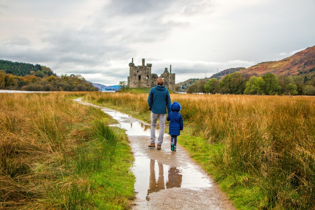 A man and a child holding hands walking along a muddy path towards a ruined castle 