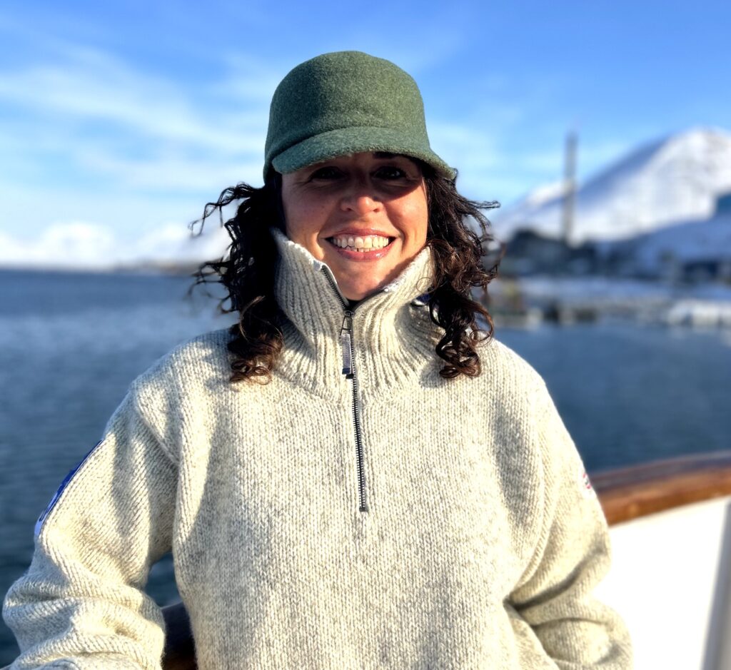 Shannon Guihan smiles from the deck of a boat, wearing a cream wool sweater with the sea in the background