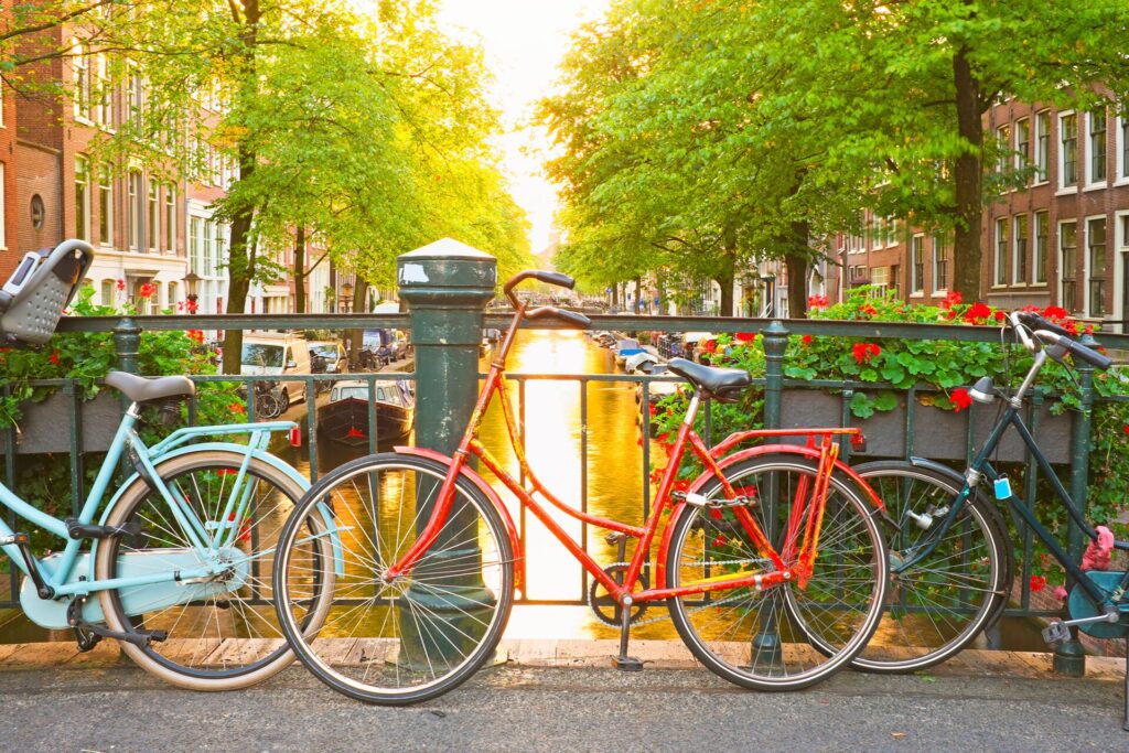 An orange bicycle rests against a railing over one of Amsterdams canals, with green trees and sunlight reflecting on the water