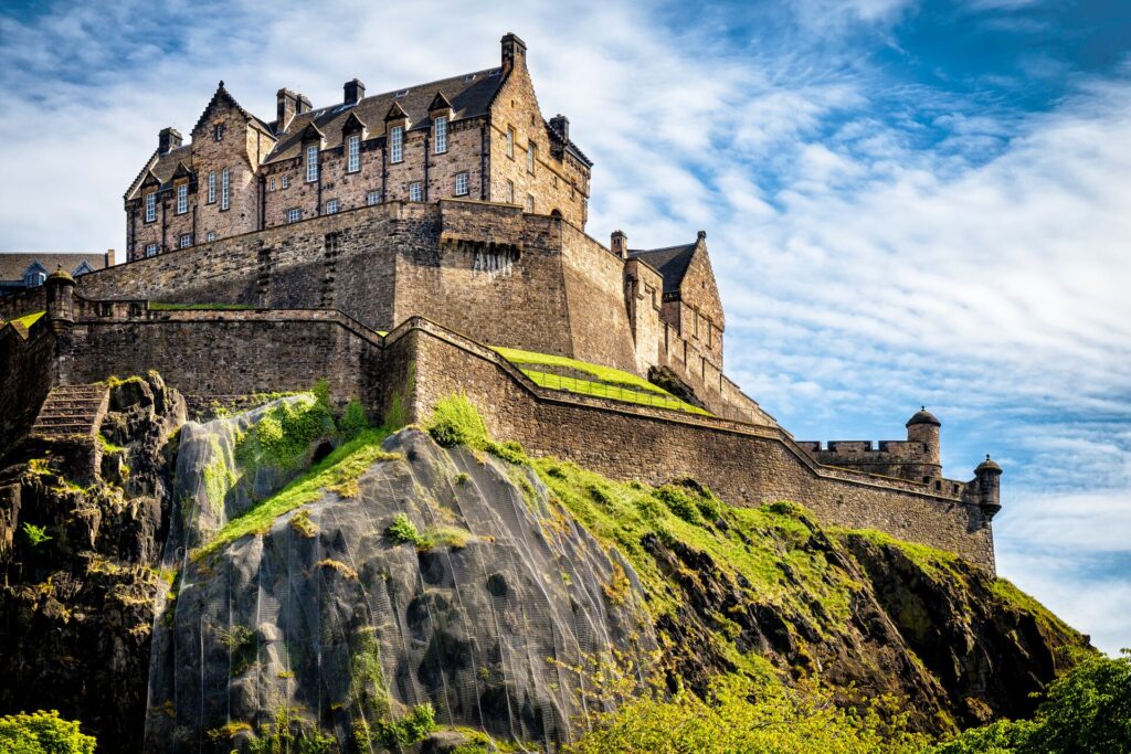The stone walls of Edinburgh Caste sit atop a grass covered tock, against a bright sky with white clouds