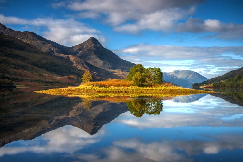 Blue skies, white clouds, a rugged mountain and foreground trees are perfectly reflected in a Scottish lake