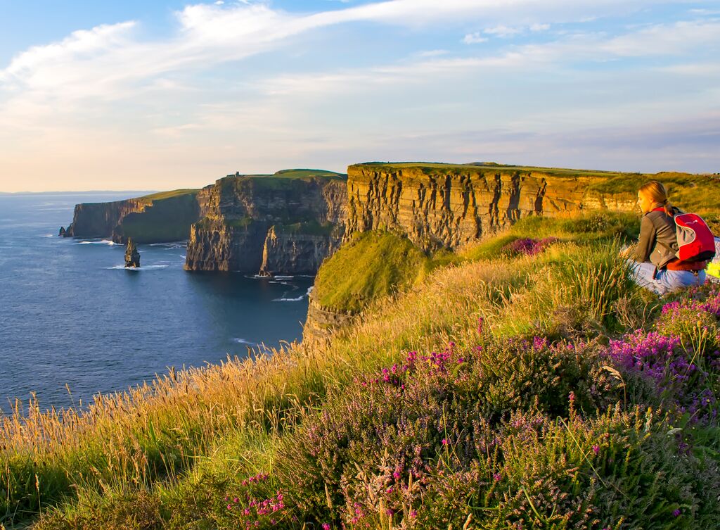 Woman looking out over cliffs and the Atlantic ocean. Wildflowers of purple and magenta cover the cliff in the foreground.
