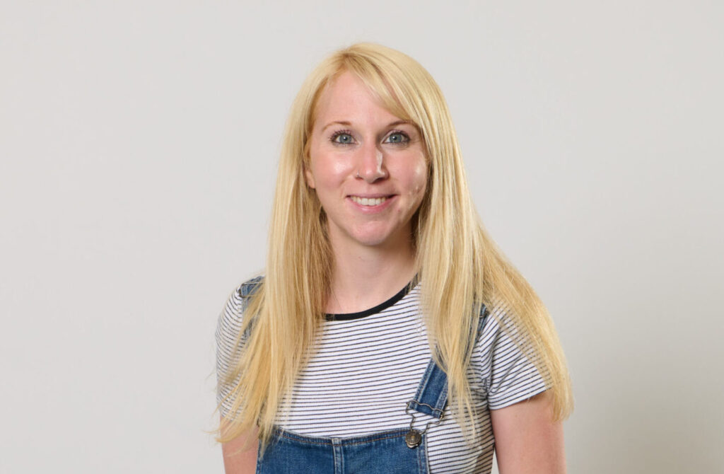 Portrait photo of Emma Harrison, wearing blue denim dungarees, standing against a white wall