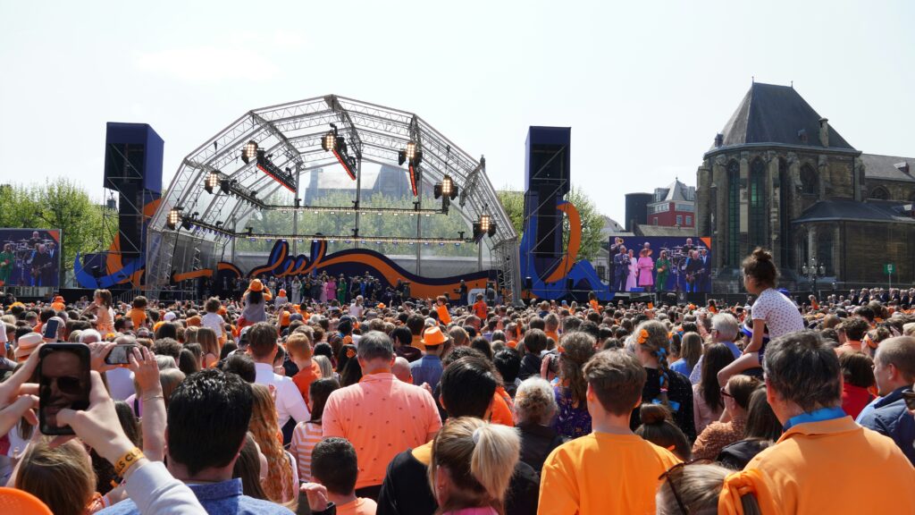 Crowds, many dressed in orange, gather in front of a stage in which the Dutch monarchy stand on Kings Day