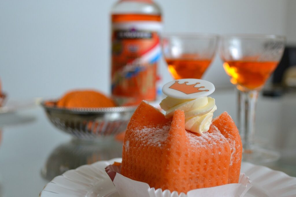 A bright orange Kings Day pastry sits in front of tow orange drinks and some orange treats on a white table