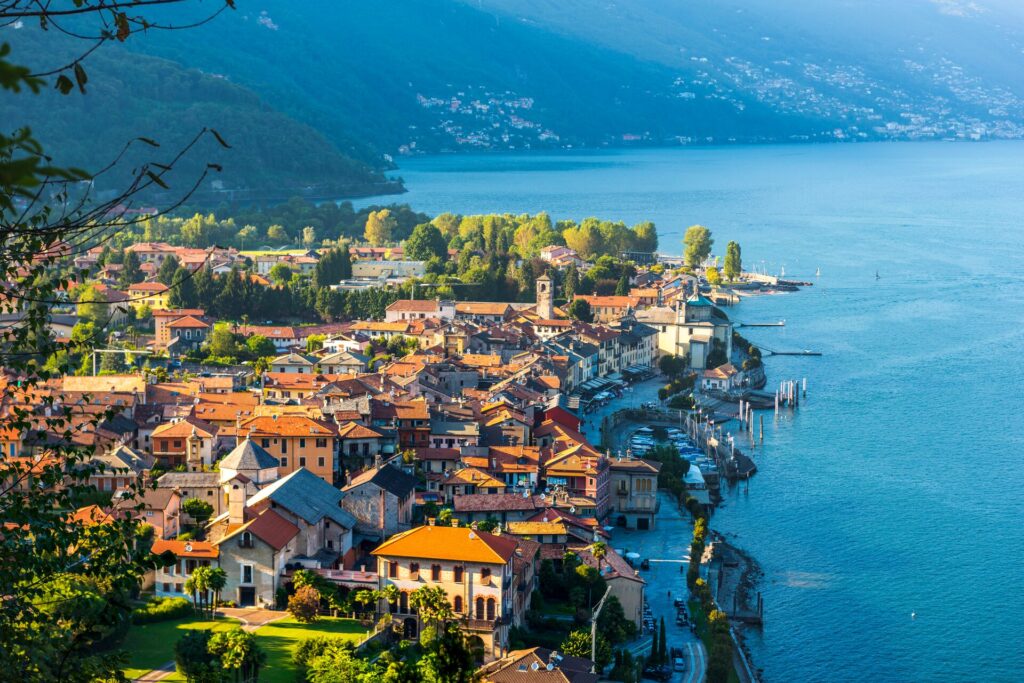 Aerial view of houses on the banks of Lake Maggiore with the lake to the left and green mountains rising up in the background. 
