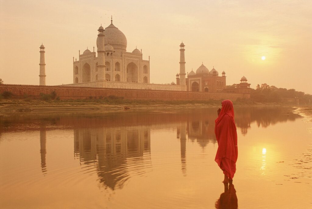 A lady draped in red standing on the banks of the river in front of the grand, white Taj Mahal in India