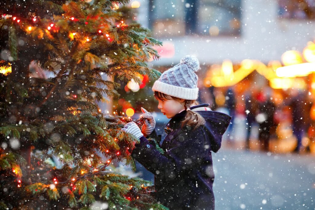 A girl looks closely at a Christmas tree as snow falls around her